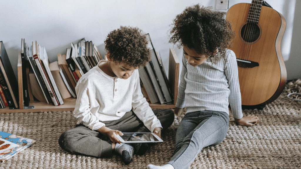 Niño y niña leyendo en una tablet, detrás hay libros y una guitarra